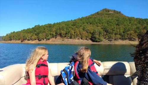 Two girls in life jackets sit on a boat, looking at a green hillside across the water on a sunny day.