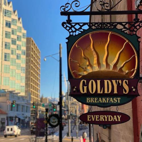 Sign for Goldy's Breakfast with a sun design, hanging outside in a city setting with buildings in the background.