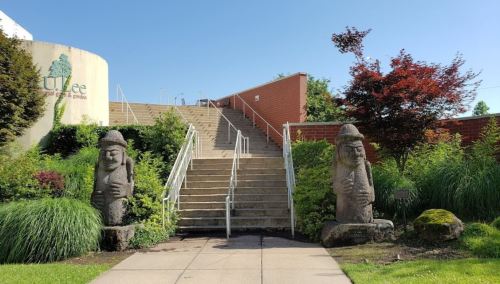 Two stone statues flank a set of stairs leading up to a building, surrounded by greenery and bright blue skies.