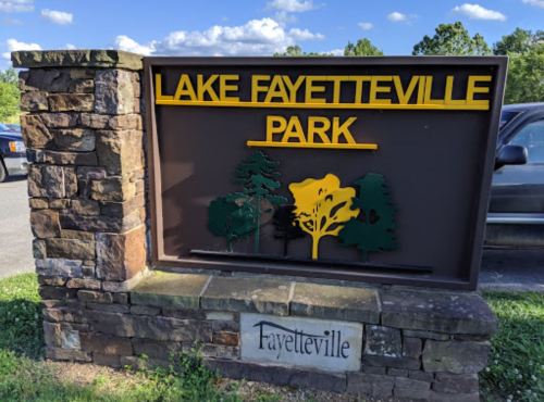 Sign for Lake Fayetteville Park featuring trees and the park name in yellow letters against a brown background.
