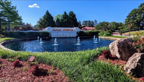 A serene pond with fountains, surrounded by greenery and flowers, featuring a sign that reads "Magic Springs."