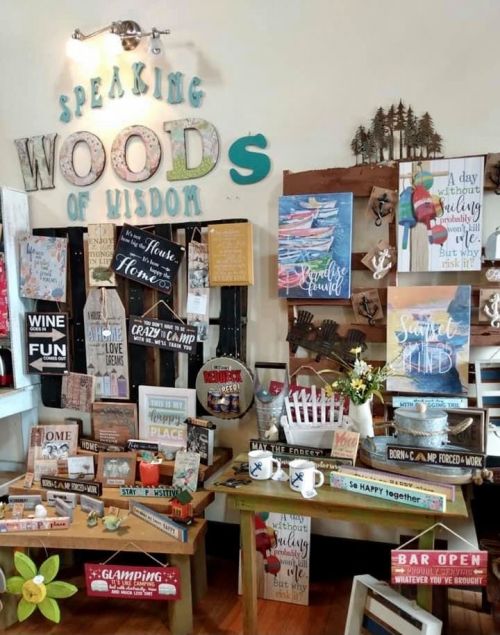 A display of wooden signs and decor with phrases about nature, camping, and home, set in a cozy shop environment.