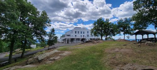 A white building on a grassy hill, surrounded by trees and under a partly cloudy sky.
