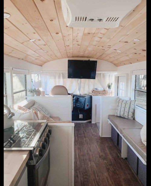 Interior of a converted bus featuring a wooden ceiling, kitchen area, seating, and a TV mounted on the wall.