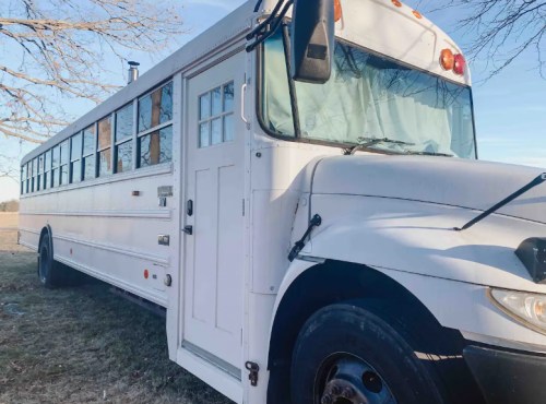 A white school bus parked on grass, with a clear sky and trees in the background.