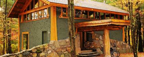 A modern cabin with wooden accents, surrounded by trees and autumn foliage, featuring a stone entrance and a bicycle.