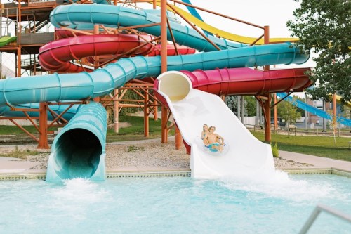 Two people slide down a white water slide at a water park, surrounded by colorful slides and a pool.