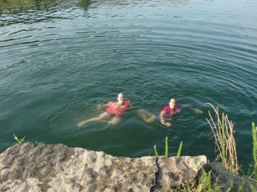 Two people swimming in a calm body of water, with a rocky shore and greenery in the background.