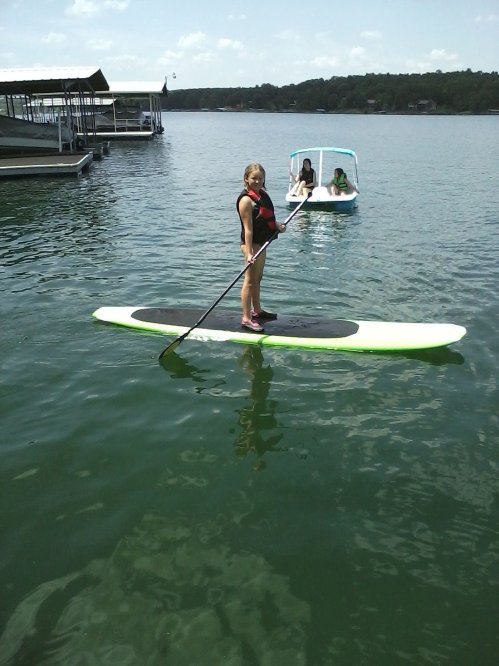 A person stands on a paddleboard in a lake, with two others in a small boat nearby. Sunny day with clear water.