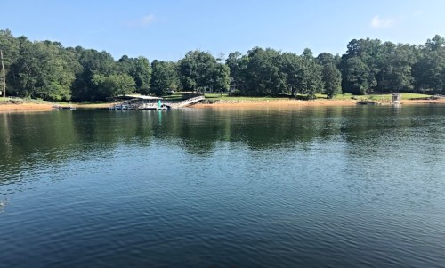 A calm lake surrounded by trees, with a dock and a sandy shore under a clear blue sky.