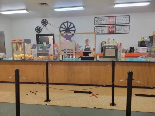 A concession stand with a smiling attendant, popcorn machine, and various snacks, featuring a colorful menu board.