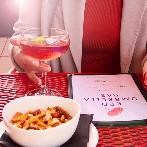 A hand holding a cocktail beside a bowl of mixed nuts and a menu on a red table.