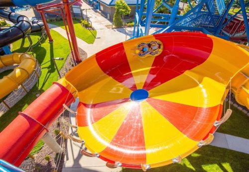 A colorful water slide with red and yellow patterns, surrounded by green grass and other slides in a water park.