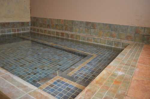 A tiled indoor pool with blue and brown mosaic patterns, featuring a shallow area and a smooth stone border.