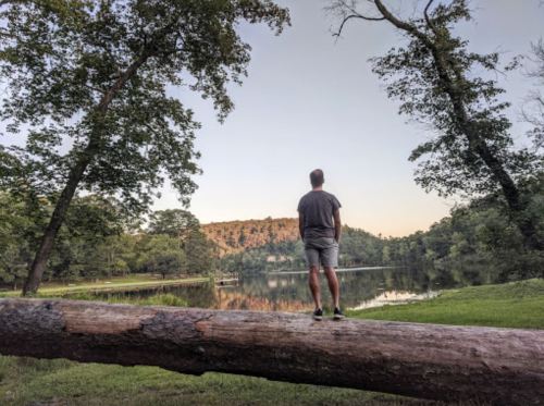 A person stands on a log by a calm lake, surrounded by trees and a scenic landscape at sunset.