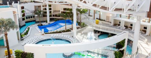 A modern resort pool area featuring multiple pools, palm trees, and lounge chairs under a bright, open structure.