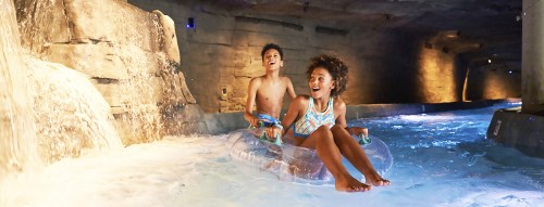 Two children enjoy a water slide in a cave-like setting, splashing through the water with smiles on their faces.
