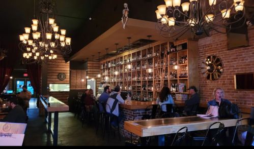 A cozy bar interior with a long wooden counter, shelves of bottles, and patrons seated at tables under chandeliers.