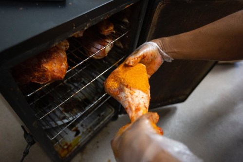 A hand wearing a glove holds a seasoned chicken leg, with more chicken pieces visible in an oven.