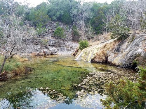 A serene natural landscape featuring a clear pond surrounded by rocky terrain and sparse vegetation.