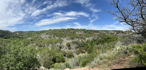 Panoramic view of a lush green landscape under a blue sky with scattered clouds and distant hills.