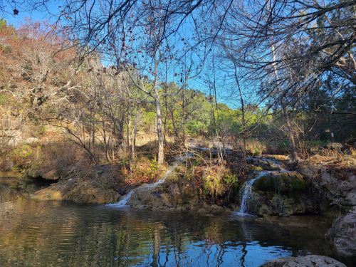 A serene landscape featuring a small waterfall flowing into a calm pool, surrounded by trees and autumn foliage.