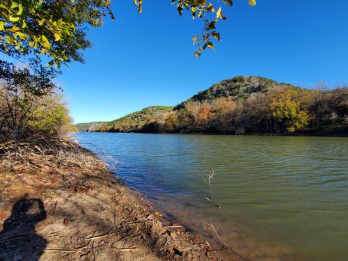 A serene river scene with lush green hills and autumn foliage under a clear blue sky. Shadow of a person in the foreground.