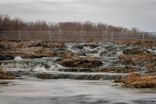 A serene river flows over rocky terrain, with sparse grass and a distant tree line under a cloudy sky.