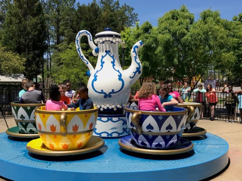Children enjoy a colorful teacup ride at an amusement park, with a large teapot centerpiece and trees in the background.