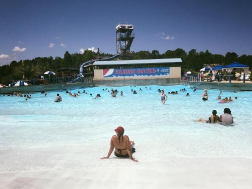 A busy water park wave pool with people swimming and lounging under a clear blue sky.
