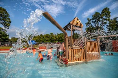 Children play in a water park pool with a large water feature and splash area under a blue sky.