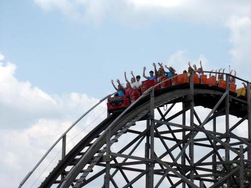 A group of excited riders on a roller coaster, raising their hands as they crest a hill against a blue sky.