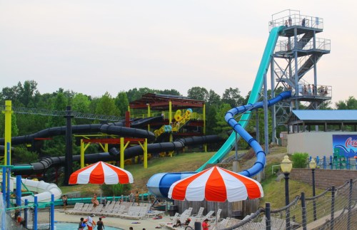 A water park scene featuring colorful slides, lounge chairs, and striped umbrellas under a cloudy sky.