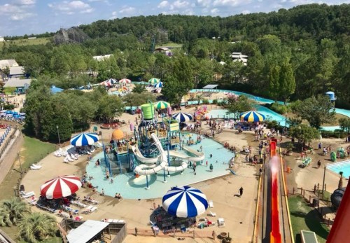 Aerial view of a vibrant water park with slides, pools, and colorful umbrellas, surrounded by trees and attractions.
