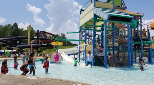 A busy water park scene with children playing in a pool, colorful slides, and a large play structure under a blue sky.