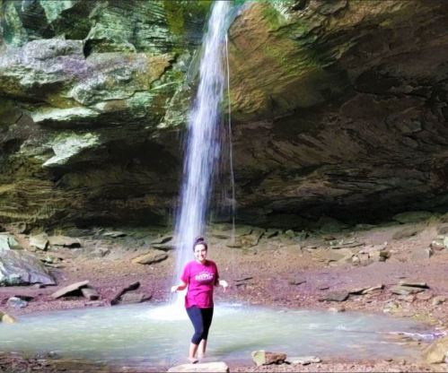 A woman stands in a shallow pool under a waterfall in a rocky cave, surrounded by lush greenery.