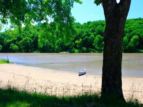 A serene river scene with a sandy shore, green trees, and a small boat moored on the water.