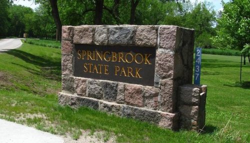 Sign for Springbrook State Park, made of stone, surrounded by green grass and trees, with a road in the background.