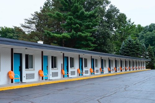A row of motel rooms with bright blue doors and orange chairs, surrounded by green trees and a paved parking area.