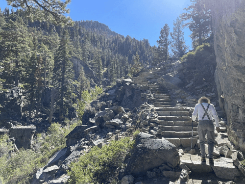 A hiker ascends stone steps through a forested mountain trail under a clear blue sky.