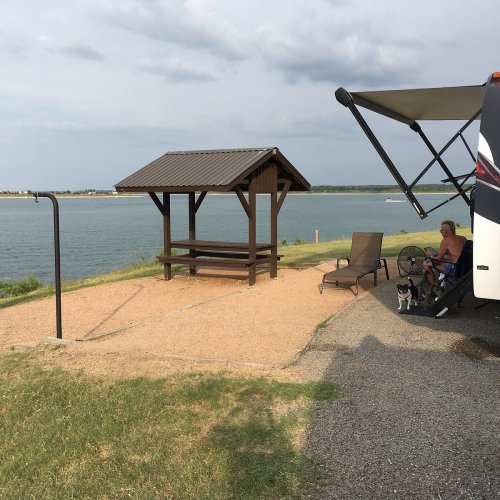 A peaceful lakeside scene with a gazebo, a person relaxing in a chair, and a dog near a camper.