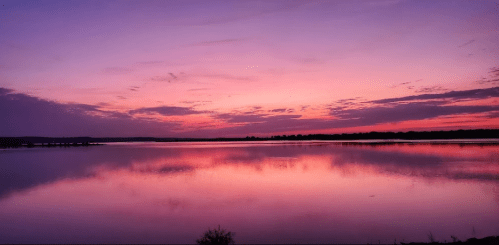 A serene lake at sunset, reflecting vibrant purple and pink hues in the sky and water.