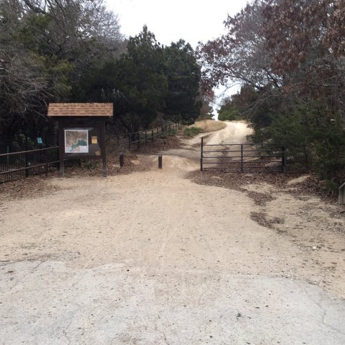 A dirt path leads to a gate, with a map sign on the left and trees lining the area. Overcast sky above.