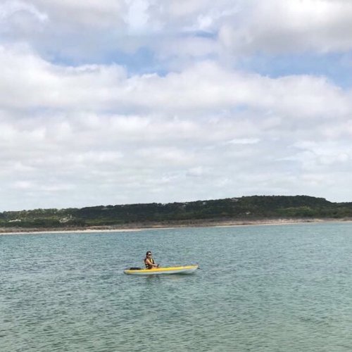 A person kayaking on calm water with a scenic shoreline and cloudy sky in the background.