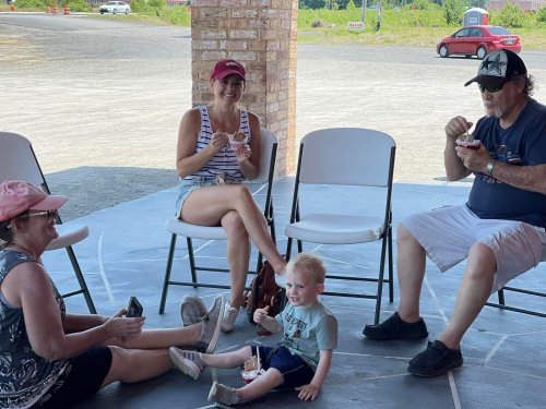 A family enjoys ice cream together, sitting on chairs and the floor in a shaded outdoor area.