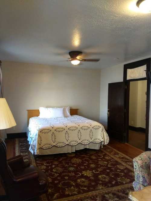 Cozy bedroom with a queen bed, ceiling fan, and a vintage armchair, featuring a patterned rug and neutral walls.