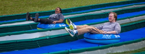 Two people joyfully sliding down a grassy slope on inflatable tubes, enjoying a fun outdoor activity.