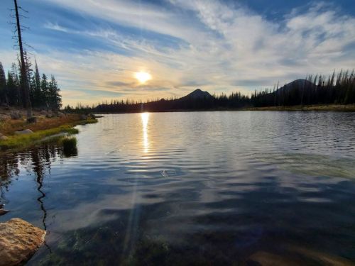 A serene lake at sunset, reflecting the sky and mountains, surrounded by trees and a peaceful landscape.