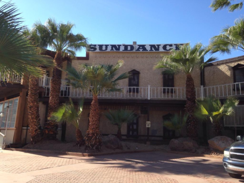 A building labeled "Sundance" surrounded by palm trees and rocky landscaping under a clear blue sky.