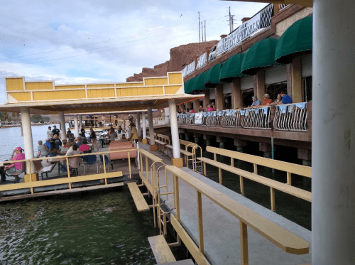 A waterfront scene with a dock, people dining on a patio, and a building with green awnings along the shore.
